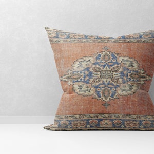May include: A decorative pillow with a vintage-style rug pattern. The pillow features a large, intricate floral design in shades of blue, red, and cream on a rust-colored background. The pillow has a fringed border.