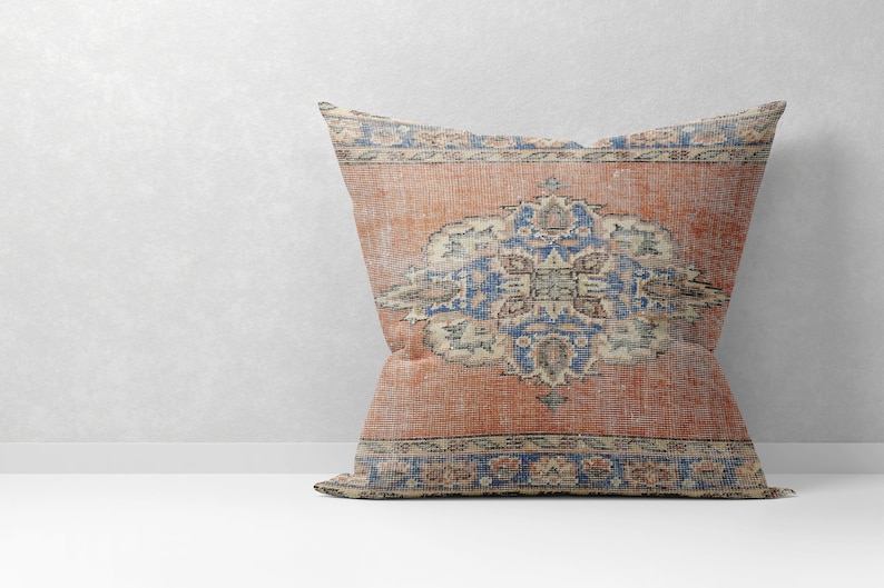 May include: A decorative pillow with a vintage-style rug pattern. The pillow features a large, intricate floral design in shades of blue, red, and cream on a rust-colored background. The pillow has a fringed border.