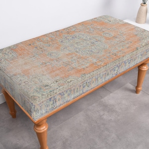 May include: A wooden bench with a patterned fabric seat. The fabric is a light orange with a blue and green floral design. The bench has four legs and is sitting on a light grey floor.