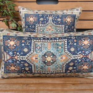 May include: Two rectangular throw pillows with a detailed, ornate pattern. The design features a central medallion in shades of orange, teal, and cream, set against a deep blue background. The pillows are placed on a wooden surface, suggesting a home decor theme.