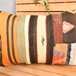 Puede incluir: Cojín rectangular decorativo con diseño de patchwork. El cojín presenta una mezcla de colores, incluyendo naranja, marrón, beige y toques de rosa y azul. El cojín está colocado sobre un banco de madera.