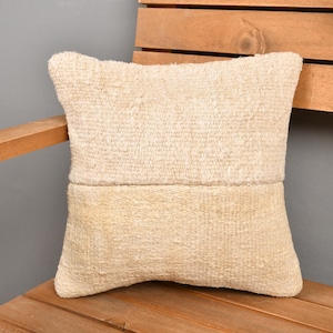 May include: A square, beige decorative pillow with a textured, woven appearance. The pillow is divided into two sections, with a slightly darker shade on the bottom half. It rests on a wooden surface.