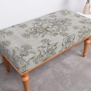 Puede incluir: Un banco gris claro con tapicería de estampado floral y patas de madera. El banco está sobre un suelo de baldosas grises.