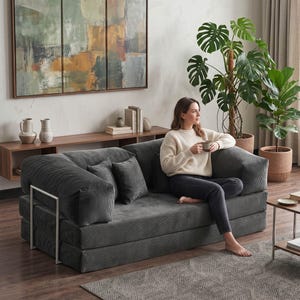 May include: A contemporary living room with a grey corduroy sofa and cushions. A woman is seated on the sofa, holding a mug. An abstract painting hangs above a wooden shelf. A large potted plant adds a natural element to the room.
