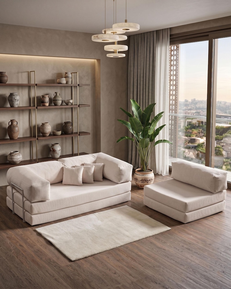 May include: A modern living room setup featuring a beige sofa, armchair, and rug. The room includes a large window, a potted plant, and a decorative shelf with vases. The furniture is arranged on a dark wood floor, creating a warm and inviting atmosphere.