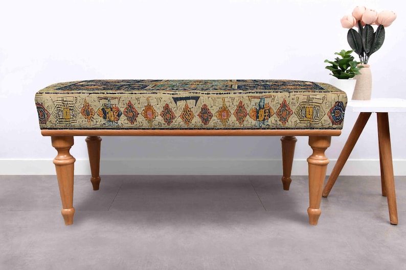 May include: A rectangular bench with a patterned top and wooden legs. The top features a beige background with a colourful, geometric design. The bench has four turned wooden legs, and is set against a white wall.