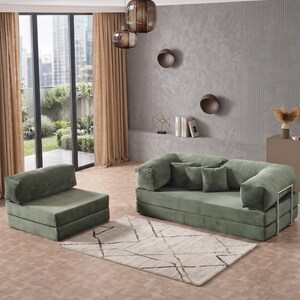 May include: A modern living room setup featuring a green sofa and armchair set with a matching rug. The sofa has a metal frame and several throw pillows. The room includes a window with tan curtains and decorative lighting.