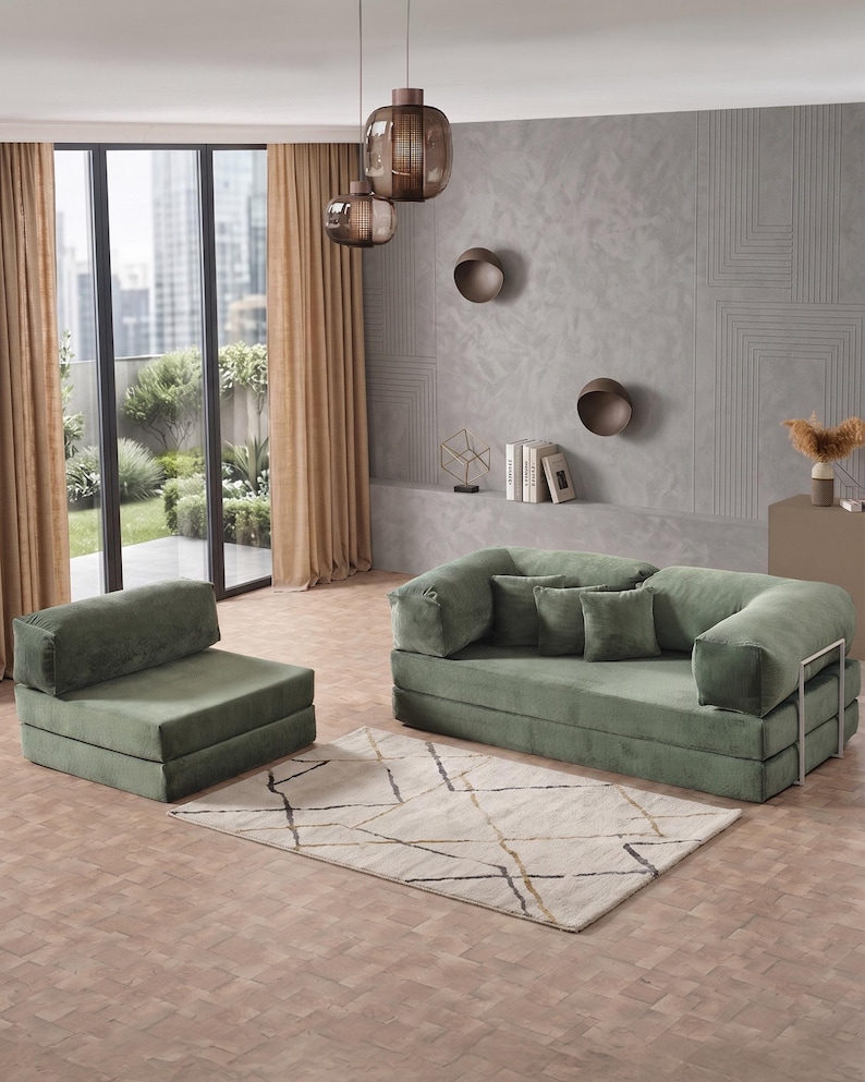 May include: A modern living room setup featuring a green sofa and armchair set with a matching rug. The sofa has a metal frame and several throw pillows. The room includes a window with tan curtains and decorative lighting.