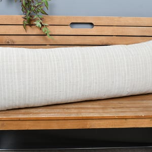 May include: A long, rectangular lumbar pillow with a light beige, vertically striped pattern. The pillow is placed on a wooden bench with a natural finish. Greenery is visible in the background.