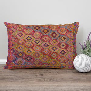 Puede incluir: Un cojín rectangular con un vibrante patrón geométrico en rojo, amarillo, azul y morado. El cojín está colocado sobre una superficie de madera, junto a una pequeña planta de lavanda en maceta y una bola decorativa.