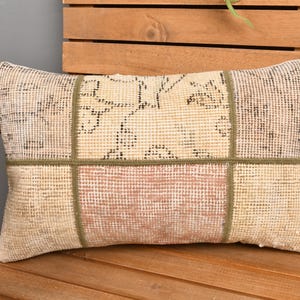 May include: Rectangular decorative pillow with a patchwork design. The pillow features a beige background with sections of light pink and patterned beige. Green stitching outlines the sections. The pillow is placed on a wooden bench.