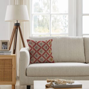 Puede incluir: Un cojín decorativo con un patrón geométrico rojo y beige y detalles florales descansa sobre un sofá beige. El cojín añade un toque de color a un salón de tonos neutros. También se ven una lámpara de pie de madera y una mesa auxiliar.