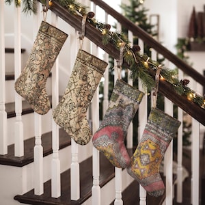 Könnte beinhalten: Vier Weihnachtsstrümpfe hängen an einem Treppengeländer aus Holz. Jeder Strumpf zeigt ein einzigartiges, komplexes und mehrfarbiges Muster, das an Vintage-Teppichdesigns erinnert, in gedeckten Grün-, Beige-, Rosa-, Gelb- und Grautönen.