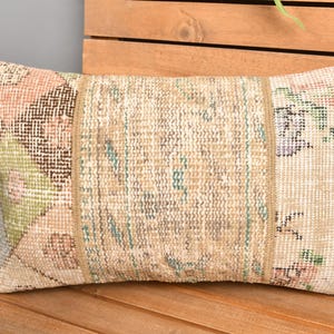 May include: Rectangular decorative pillow with a patchwork design. The pillow features a central section with vertical stripes and floral patterns on the ends. The color palette includes beige, pink, green, and brown tones. The pillow is placed on a wooden surface.
