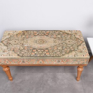 May include: A rectangular ottoman with a patterned rug top in shades of green, beige, and peach. The ottoman has four wooden legs. A small white side table with a vase of flowers and a potted plant is next to the ottoman.