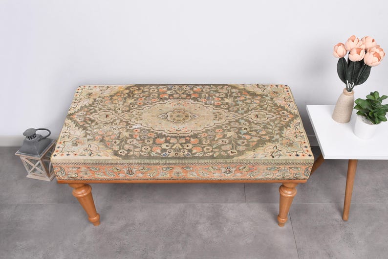May include: A rectangular ottoman with a patterned rug top in shades of green, beige, and peach. The ottoman has four wooden legs. A small white side table with a vase of flowers and a potted plant is next to the ottoman.