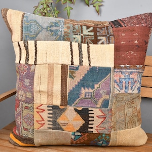 Op de afbeelding: Een vierkant patchwork kussen met een verscheidenheid aan kleuren en patronen. Het kussen heeft een mix van beige, blauwe, rode en paarse tinten, met geometrische en bloemmotieven. Het ligt op een houten bank.