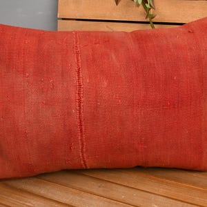May include: A rectangular, rust-colored pillow with a textured surface. The pillow is placed on a wooden bench. The pillow has a seam down the center and is made of a woven fabric.