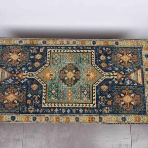 May include: A rectangular rug with a detailed geometric pattern. The rug has a dark blue base with teal, orange, and yellow elements. The design includes a central medallion and border details.