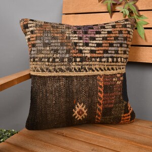 Op de afbeelding: Vierkant decoratief kussen met een patchwork ontwerp. Het kussen heeft een mix van bruine, zwarte, oranje en beige tinten. Het bovenste gedeelte heeft een geweven patroon, terwijl de onderkant een donkerdere, getextureerde uitstraling heeft. Een uniek woondecoratie-item.