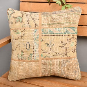May include: Square beige pillow with a patchwork design. The pillow features rectangular sections with faded patterns in shades of beige, teal, and brown. The pillow is placed on a wooden surface, suggesting a rustic or bohemian style.