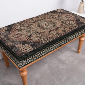 May include: A wooden bench with a patterned cushion. The cushion has a black, green, and brown geometric design. The bench has four wooden legs.