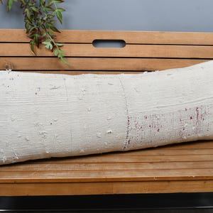 May include: A long, rectangular off-white cushion with a textured surface and subtle red accents. The cushion is placed on a wooden bench, suggesting a rustic or bohemian style. The cushion's design is simple and likely suitable for a variety of decor styles.