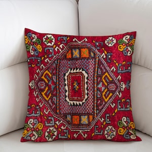 May include: A decorative square pillow with a vibrant red background and a detailed geometric pattern in shades of purple, yellow, blue, and orange. The pillow is placed on a white sofa, showcasing its intricate design and color palette.