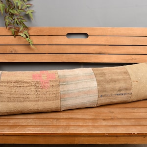 Op de afbeelding: Een lang, rechthoekig kussen met een patchworkontwerp in tinten beige, bruin en roze. Het kussen ligt op een houten bank. De textuur van het kussen lijkt geweven te zijn, met zichtbare naden en variaties in de stof.