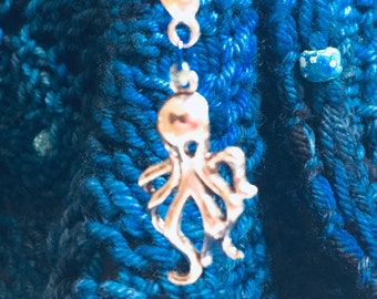 May include: A silver octopus charm hangs from a lobster clasp attached to a red cord. The charm is suspended against a backdrop of blue and teal knitted fabric. A small blue and white bead is also visible.