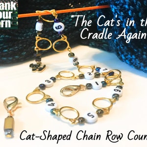 May include: A gold cat-shaped chain row counter with black and white beads and gold rings. The counter is attached to a knitting needle with a quote that reads "The Cat's in the Cradle Again".