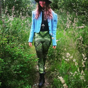 May include: A woman wearing a light blue jacket, a brown hat, and green patterned leggings stands in a field of green grass and wildflowers.
