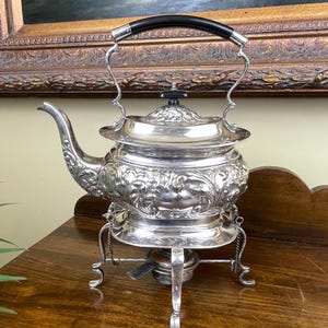 May include: A silver tea kettle with ornate floral detailing, a black handle, and a warming stand. The kettle has a spout and a lid with a small black knob. The stand has three legs and a small burner. The kettle is sitting on a wooden surface.