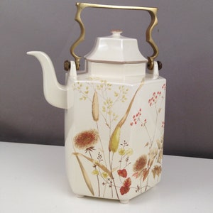 May include: Cream-colored ceramic teapot with a brass handle and lid. The teapot features a floral design with brown, yellow, and red accents. The spout is curved, and the handle is ornate. The teapot is a vintage style.