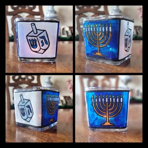 May include: Four square glass candle holders with blue and white stained glass designs. Two of the holders feature a menorah with gold candles and the other two feature a dreidel with the Hebrew letter 'nun' on it.