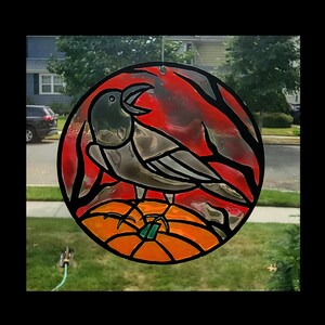 May include: A round stained glass window decoration featuring a crow perched on an orange pumpkin. The crow is gray and black, set against a red and black background. The pumpkin has a green stem. The window is hanging in front of a window.
