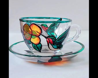 May include: A glass teacup and saucer with a hand-painted design of a hummingbird and a flower. The hummingbird is green, red, and black, and the flower is yellow, orange, and green. The cup and saucer have a green rim.