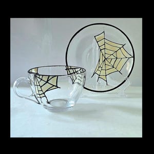 May include: A clear glass teacup and saucer set. The teacup has a handle and a spiderweb design in black and cream. The matching saucer also features a spiderweb design. The set is displayed on a white surface.
