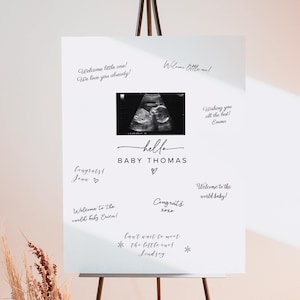 Puede incluir: Un letrero blanco sobre un caballete negro con el texto "hello BABY THOMAS" y una imagen de ultrasonido. El letrero está rodeado de mensajes escritos a mano. Un jarrón con flores secas y una hoja de palma está frente al caballete.