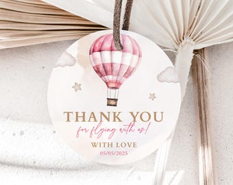 May include: A round paper gift tag featuring a watercolor pink and white hot air balloon, clouds, and stars. The text reads: THANK YOU for flying with us! WITH LOVE 05/05/2025. The tag includes a brown leather cord for hanging.