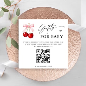 Może przedstawiać: Biała karta z napisem "Prezenty dla dziecka" i kodem QR, ozdobiona akwarelowymi wiśniami i różową kokardą. Karta znajduje się na powierzchni w kolorze różowego złota, z białą wstążką i zielenią w tle.