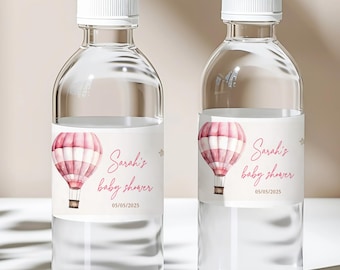 Puede incluir: Dos botellas de agua de plástico transparente con tapones de rosca blancos, cada una con una etiqueta blanca que muestra una ilustración de globo aerostático en acuarela rosa, el texto Sarah's baby shower y la fecha 05/05/2025.
