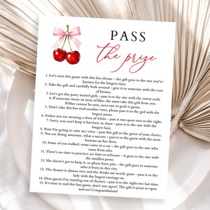 May include: A white card with the text "PASS the prize" in red script, featuring a cherry illustration. The card contains instructions for a party game. The background is a neutral, textured surface.