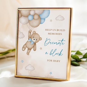 May include: A framed print with a gold border features a watercolor illustration of a teddy bear holding balloons. The text reads "Help us build memories. Decorate a block for baby."