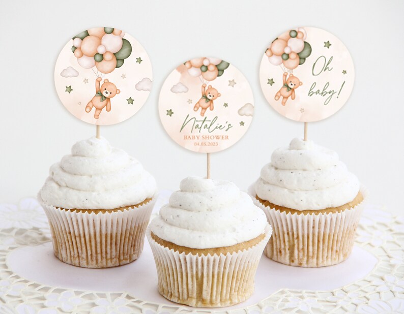 May include: Three cupcake toppers with a teddy bear holding balloons. The toppers are decorated with peach, green, and white balloons, clouds, and stars. The text on the toppers reads "Oh baby!" and "Natalie's Baby Shower 04.08.2023".
