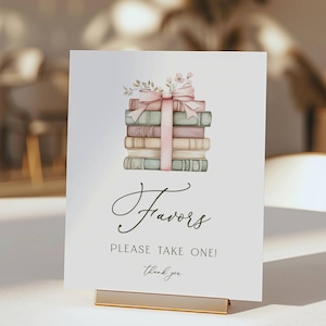 May include: A white rectangular sign featuring a watercolor illustration of a stack of books tied with a pink ribbon and small flowers. The text reads "Favors", "PLEASE TAKE ONE!", and "Thank you" in black script and sans-serif fonts.
