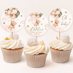 May include: Three cupcakes decorated with white frosting and topped with a small white ball and a gold star. Each cupcake has a round, white and peach-colored paper topper with a cartoon illustration of a teddy bear holding balloons. The text on the toppers reads "Oh baby!" and "Natalie's Baby Shower 04.05.2023".
