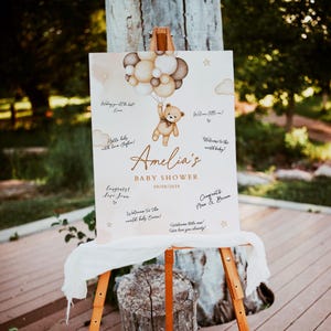 May include: A white sign with a brown teddy bear holding balloons. The sign says "Amelia's Baby Shower 09/08/2024" and has space for guests to write messages.
