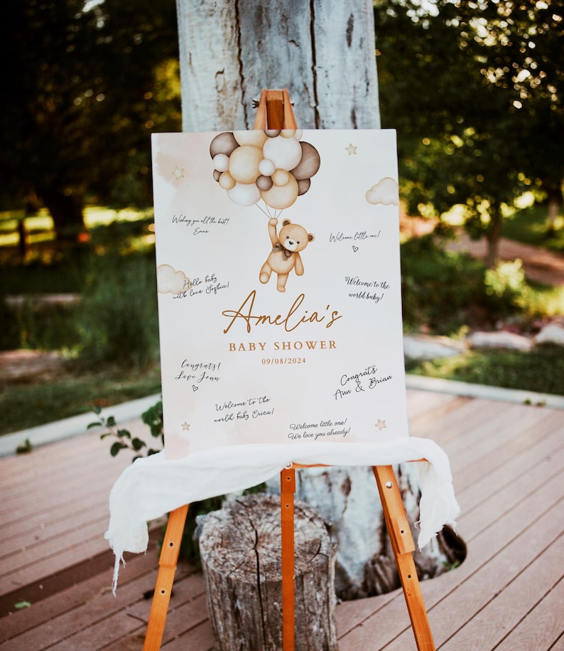 May include: A white sign with a brown teddy bear holding balloons. The sign says "Amelia's Baby Shower 09/08/2024" and has space for guests to write messages.
