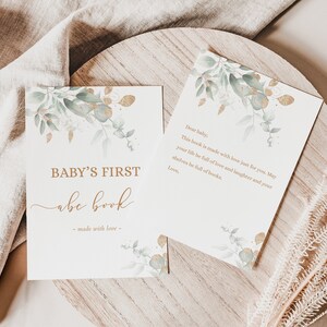 May include: A white card with gold lettering that says "BABY'S FIRST abc book - made with love -" with a watercolor illustration of green leaves and gold accents.  The card is on a wooden surface with a white cloth in the background.